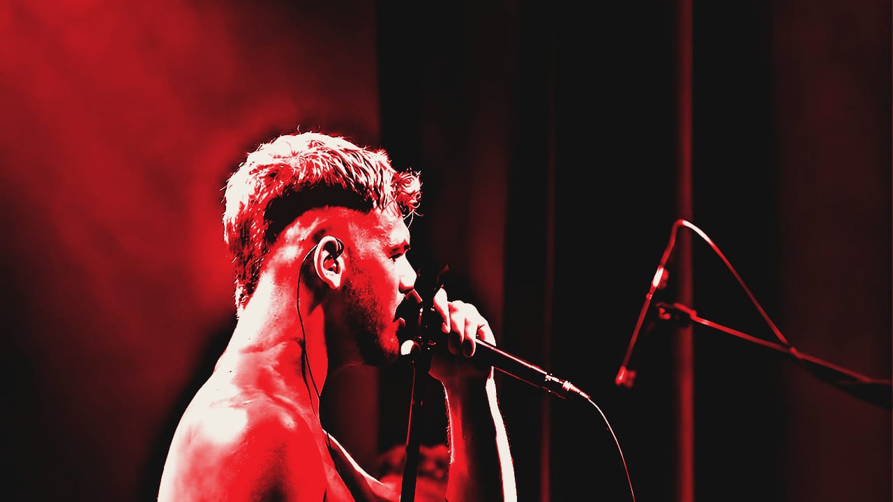 Daan, zanger van De Peppers, zingt met ontbloot bovenlijf in warm licht tegen een blauw podiumdoek